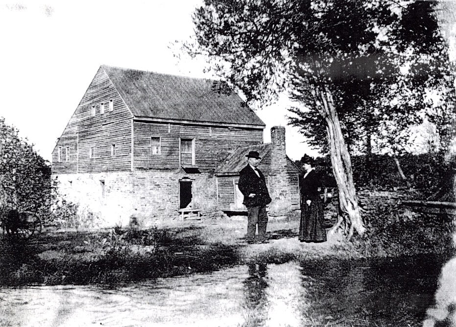 Old black & white photograph of the gristmill Miller with a woman, standing in front of the mill house.